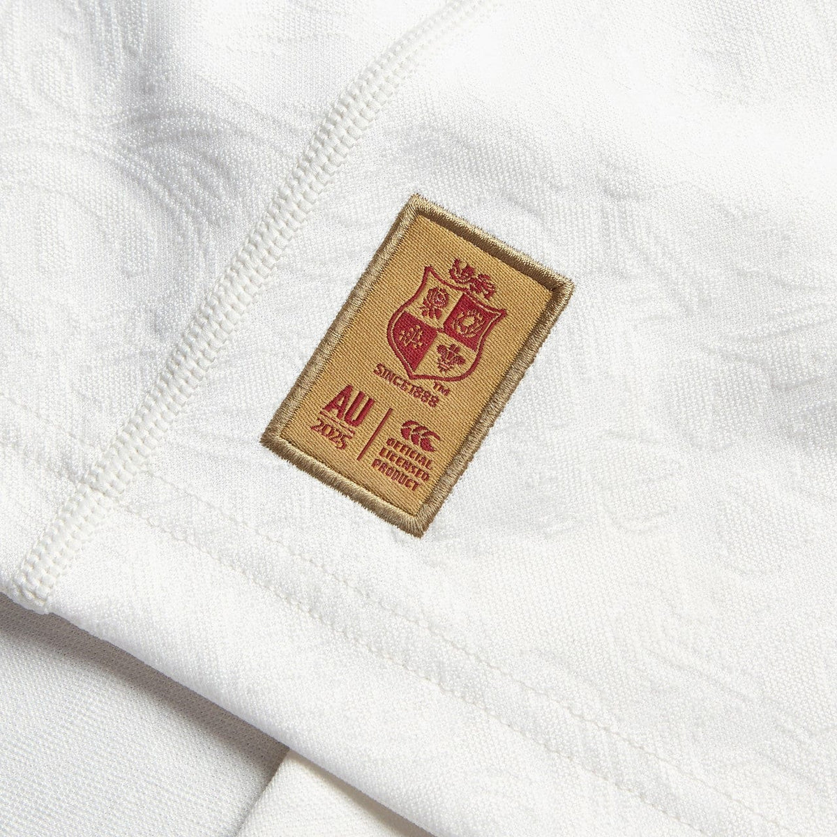 Close-up of a white Training Jersey by Canterbury, showcasing a patch with the British &amp; Irish Lions shield logo and the text "AU 2025." The patch is bordered in brown with red details, made from recycled polyester. Seams are visible around the patch.