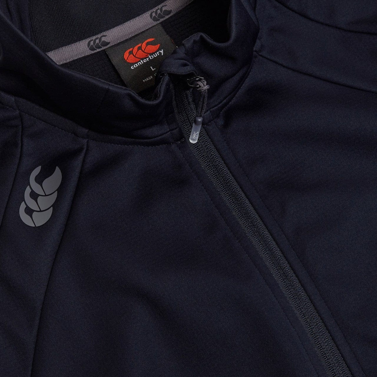 Close-up of a dark Canterbury Elite Windstopper Jacket with a Canterbury logo on the left chest and a partially unzipped front, designed for weatherproofing &amp; mobility.